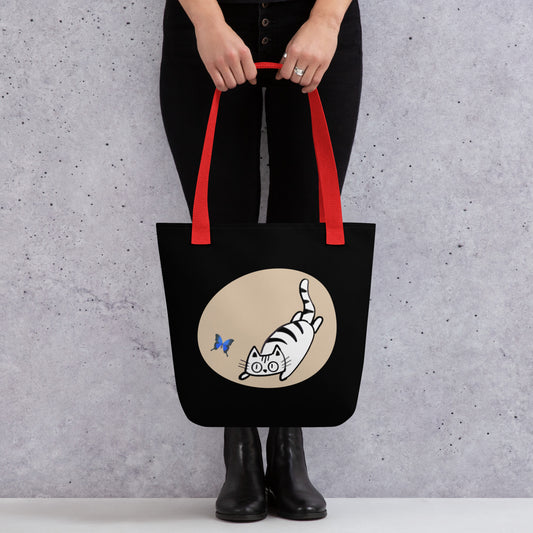 Front View of a tote bag with a design of a cat chasing a butterfly. Red Handle.
