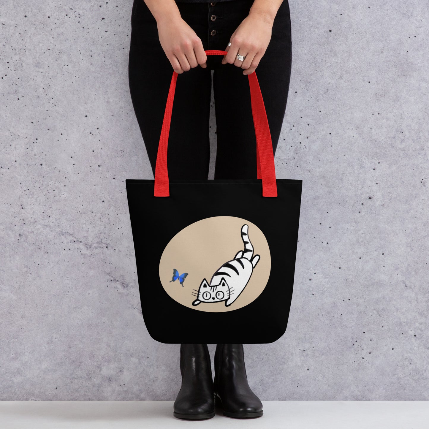 Front View of a tote bag with a design of a cat chasing a butterfly. Red Handle.
