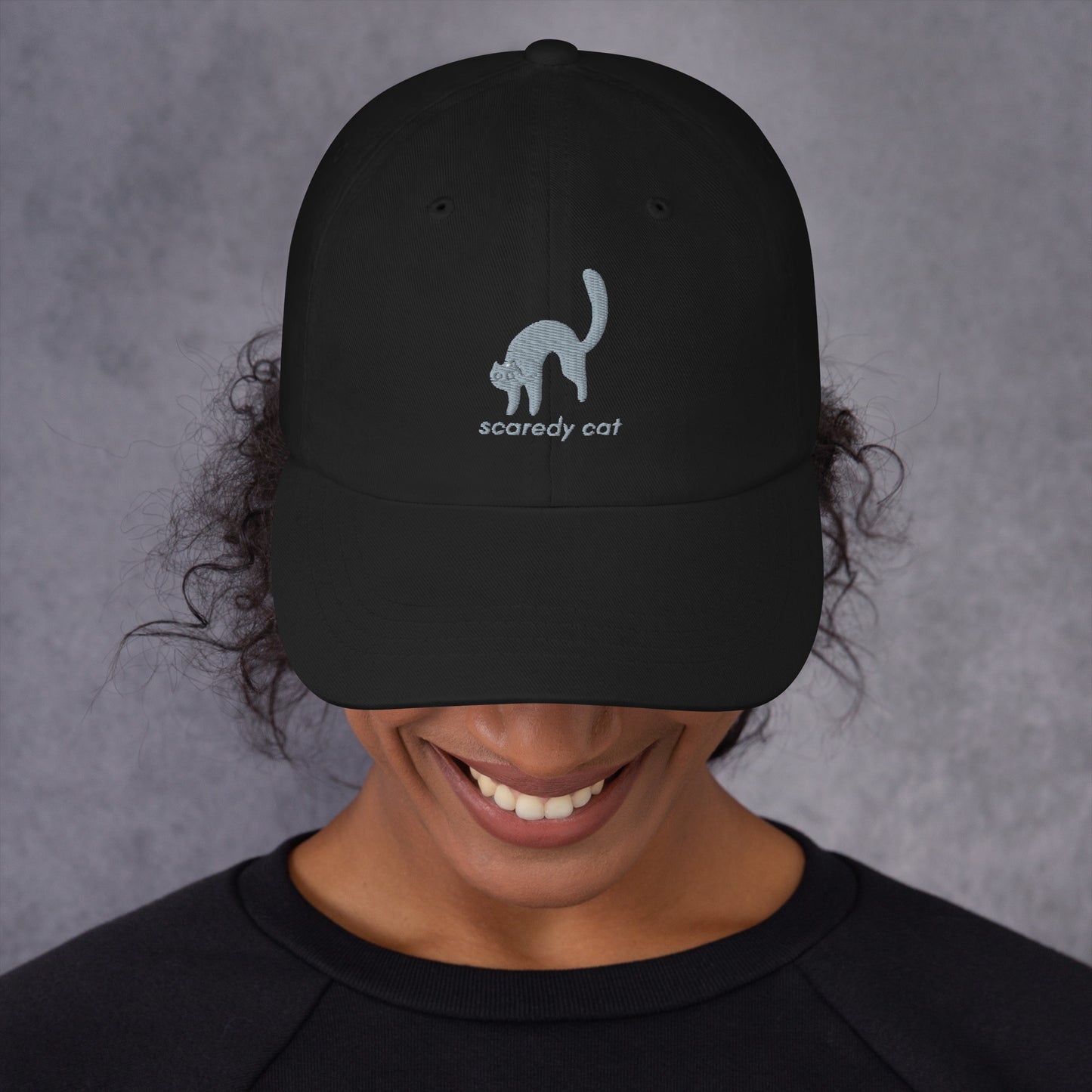 Woman wearing a black cap with a design of a scared cat with the words "Scaredy Cat".
